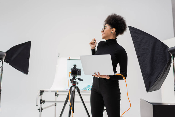 現代のフォトスタジオでソフトボックスやデジタルカメラの近くにラップトップを持つ陽気なアフリカ系アメリカ人のコンテンツメーカー - 写真・画像