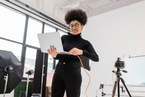 alegre afroamericano gestor de contenidos enchufar el cable en el ordenador portátil cerca de trípode con cámara digital en el estudio de fotos moderno - Foto, Imagen