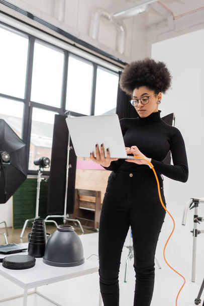アフリカ系アメリカ人のコンテンツマネージャーがノートパソコンを使いながら写真スタジオの照明器具の近くに立ち - 写真・画像
