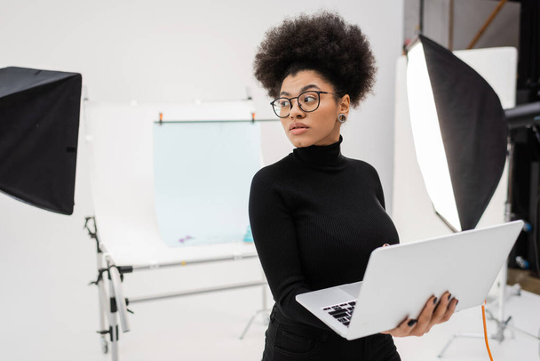 african american content manager with laptop looking away near spotlights and blurred shooting table in photo studio - Photo, Image