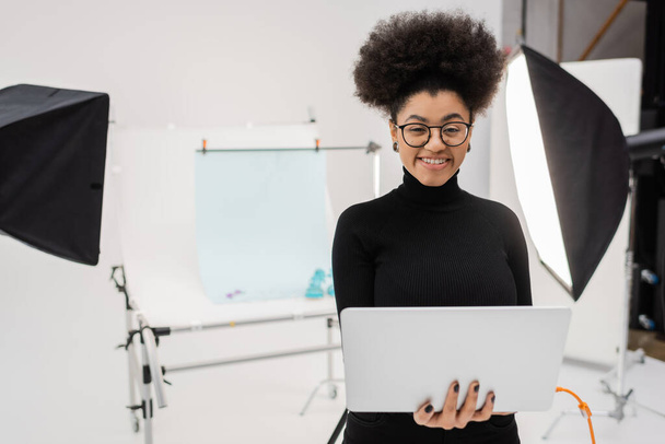 heureux producteur de contenu afro-américain avec ordinateur portable souriant à la caméra près des réflecteurs et table de tir en studio photo - Photo, image
