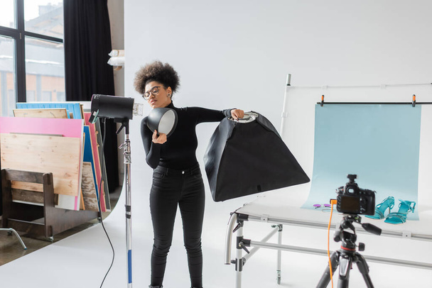 Αφροαμερικανός παραγωγός περιεχομένου που συναρμολογεί εξοπλισμό φωτισμού και ανακλαστήρα στο σύγχρονο στούντιο - Φωτογραφία, εικόνα