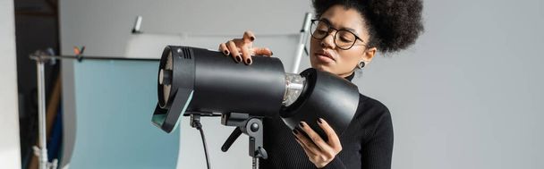 fabricant de contenu afro-américain élégant dans les lunettes de travail avec un équipement d'éclairage professionnel dans le studio photo, bannière - Photo, image