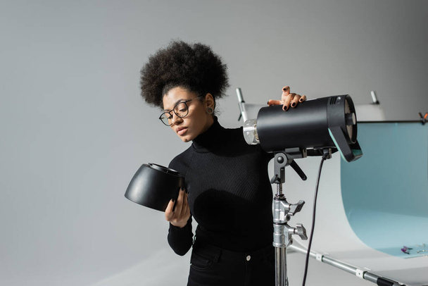 african american content maker in black turtleneck and eyeglasses holding detail of strobe lamp in photo studio - Photo, Image