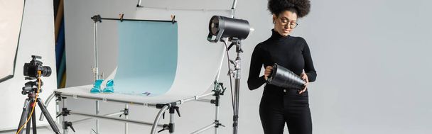 Αφροαμερικανός διευθυντής περιεχομένου που συναρμολογεί λάμπα στροβοσκοπήσεων κοντά σε ψηφιακή φωτογραφική μηχανή και τραπέζι γυρισμάτων σε φωτογραφικό στούντιο, banner - Φωτογραφία, εικόνα