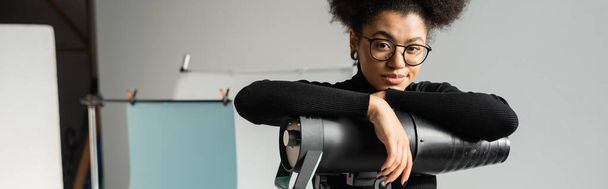 スタイリッシュなメガネでアフリカ系アメリカ人のコンテンツ制作者がスタジオのストローブランプの近くのカメラを見て - 写真・画像
