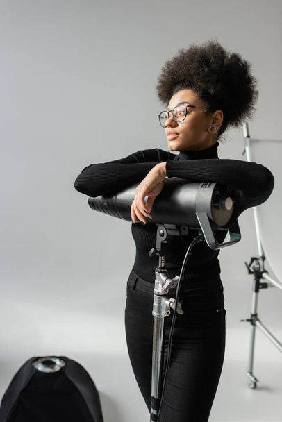 african american content producer in eyeglasses and black turtleneck looking away near strobe floodlight in photo studio - Foto, Imagem