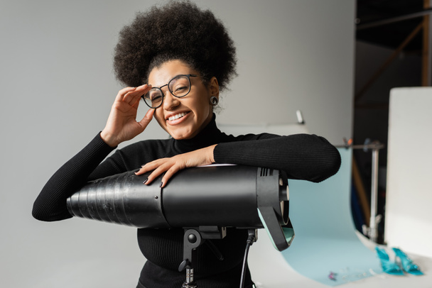 Neşeli Afro-Amerikan içerik üreticisi şık gözlükleri ayarlıyor ve fotoğraf stüdyosundaki flaş lambanın yanındaki kameraya bakıyor. - Fotoğraf, Görsel
