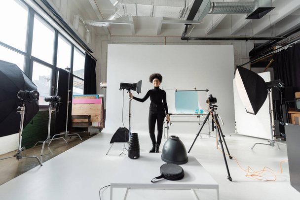 現代のスタジオのストロボランプの近くに立つ黒い服のアフリカ系アメリカ人のコンテンツマネージャーの長さ - 写真・画像