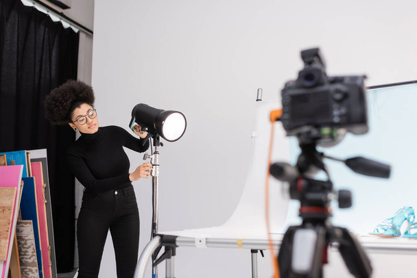 african american content maker adjusting floodlight near shooting table and blurred digital camera in photo studio - Photo, Image