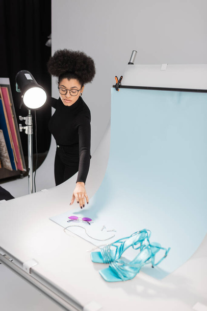 african american content producer near strobe lamp and shooting table with stylish sunglasses and sandals in photo studio - Photo, Image