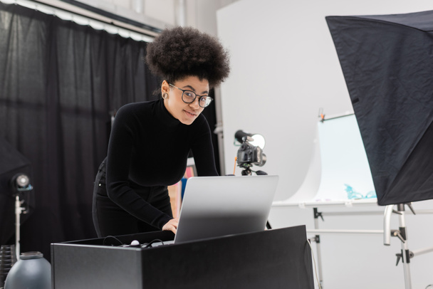 Lächelnder afrikanisch-amerikanischer Content Manager mit Brille und schwarzem Rollkragen, der im Fotostudio in die Kamera neben dem Laptop blickt - Foto, Bild
