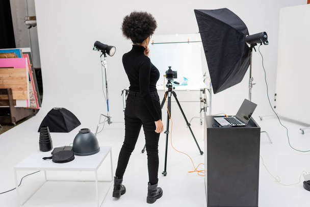 back view of african american content manager in black clothes standing near laptop and digital camera on tripod in photo studio - Φωτογραφία, εικόνα