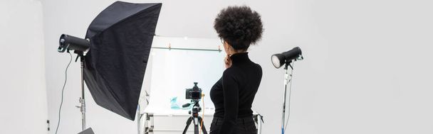 πίσω όψη της αφρικανικής Αμερικανός διευθυντής περιεχομένου σε μαύρα ρούχα κοντά σε εξοπλισμό φωτισμού και ψηφιακή φωτογραφική μηχανή στο στούντιο φωτογραφιών, banner - Φωτογραφία, εικόνα