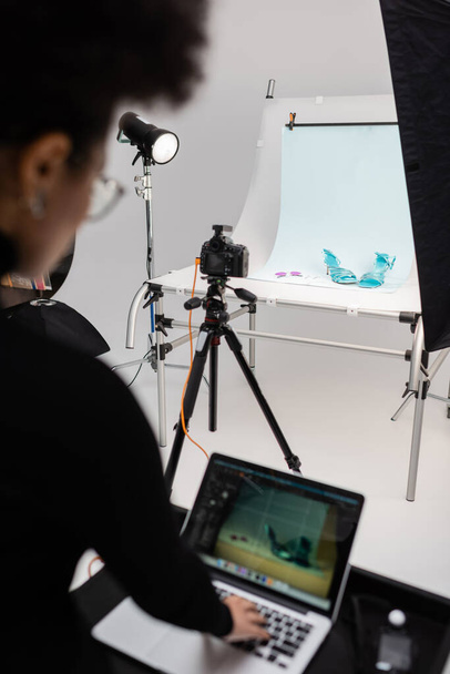 produtor de conteúdo afro-americano borrado trabalhando em laptop perto de câmera digital e calçados na moda na mesa de tiro no estúdio de fotos - Foto, Imagem