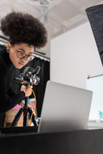 Αφροαμερικάνος παραγωγός περιεχομένου σε γυαλιά ηλίου που εργάζεται κοντά σε laptop και ψηφιακή κάμερα σε τρίποδο στο φωτογραφικό στούντιο - Φωτογραφία, εικόνα