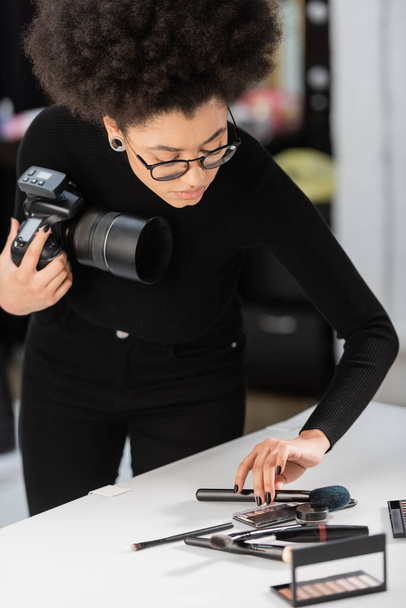 african american content producer with digital camera placing cosmetic brush near decorative cosmetics on shooting table in photo studio - Photo, Image
