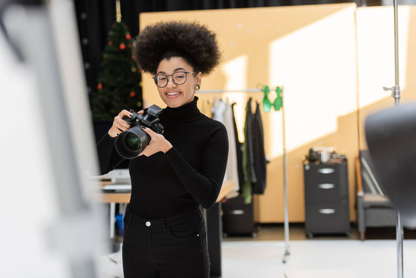 ευχαριστημένος αφρικανός Αμερικανός φωτογράφος στέκεται με επαγγελματική ψηφιακή φωτογραφική μηχανή στο στούντιο φωτογραφιών - Φωτογραφία, εικόνα
