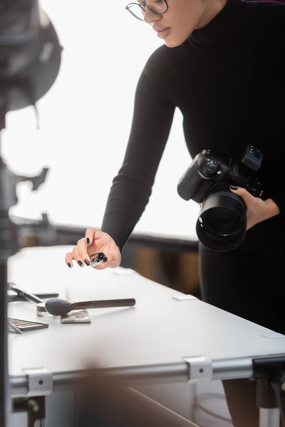 partial view of african american content producer with digital camera and mascara near decorative cosmetics on shooting table in photo studio - Foto, afbeelding