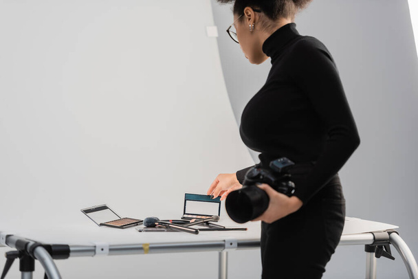 africký americký tvůrce obsahu s digitálním fotoaparátem dotýkající se oční stíny paleta v blízkosti dekorativní kosmetiky na fotografickém stole - Fotografie, Obrázek