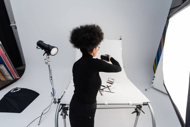 back view of african american photographer taking picture of beauty tools and decorative cosmetics on shooting table in photo studio - Photo, Image