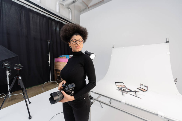 happy african american content maker with digital camera looking away near decorative cosmetics on shooting table in photo studio - Photo, Image