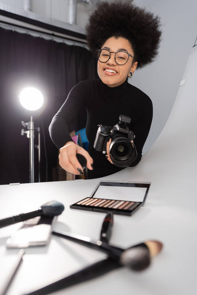 cheerful african american photographer in eyeglasses looking at camera near decorative cosmetics on shooting table in photo studio - Photo, Image
