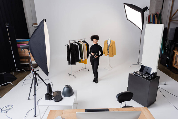 producteur de contenu afro-américain complet avec appareil photo numérique près des vêtements à la mode dans un studio photo spacieux avec équipement d'éclairage - Photo, image