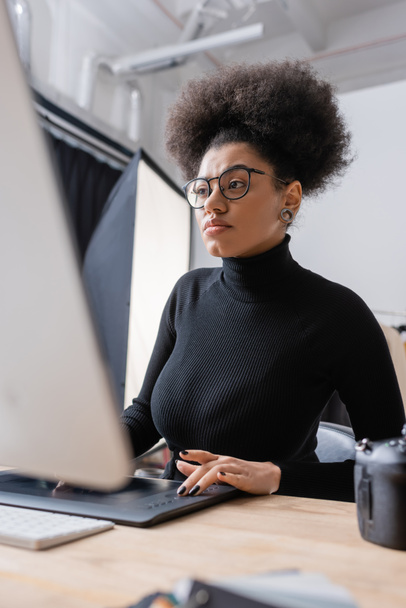 concentrated african american retoucher in black turtleneck and eyeglasses working on computer and graphic tablet in photo studio - Φωτογραφία, εικόνα
