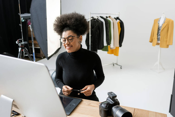onnellinen afrikkalainen amerikkalainen sisällöntuottaja katselee tietokoneen monitoria lähellä digitaalikameraa ja trendikkäitä vaatteita taustalla valokuvastudiossa - Valokuva, kuva