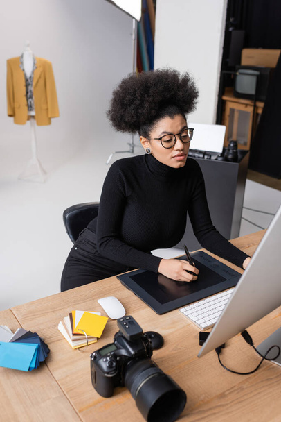 afrikanisch-amerikanische Retuschiererin in Brille arbeitet an Grafik-Tablet und Computer in der Nähe von Digitalkameras und Farbmustern im Fotostudio - Foto, Bild