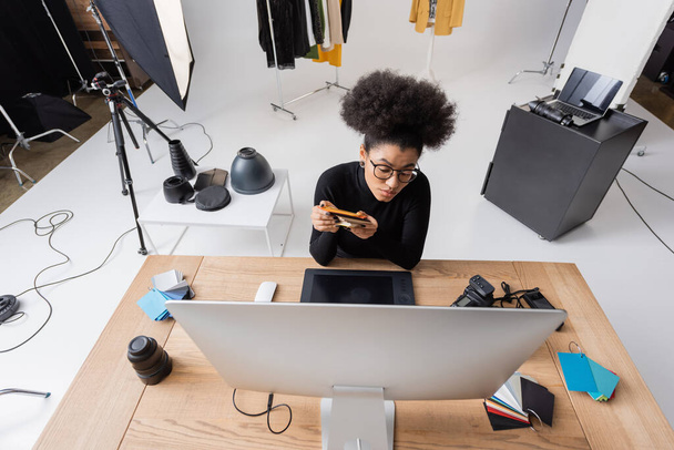 Modern fotoğraf stüdyosunda renkli örnekler ve grafik tablet yanında oturan Afro-Amerikan rötuş cihazının yüksek açılı görüntüsü - Fotoğraf, Görsel