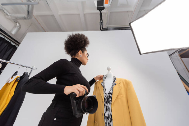 low angle view of african american photographer with digital camera adjusting stylish blazer in photo studio - Photo, Image