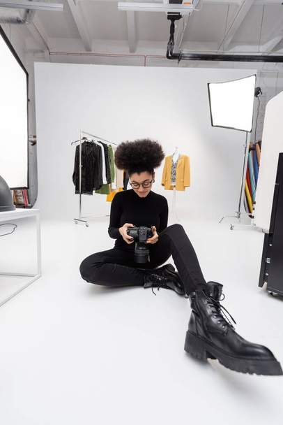 longitud completa de fabricante de contenido afroamericano feliz sentado con cámara digital cerca de focos y ropa de moda en el estudio de fotos - Foto, Imagen