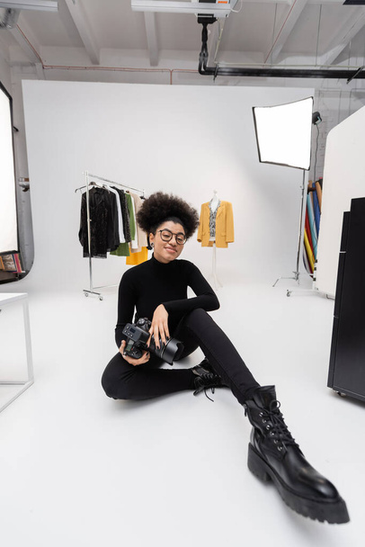 african american content producer in eyeglasses and black clothes smiling at camera while sitting in photo studio - Photo, Image