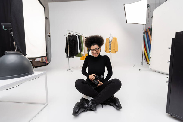 alegre afroamericano fotógrafo con cámara digital sentado con las piernas cruzadas cerca de ropa de moda en estudio de fotos - Foto, imagen