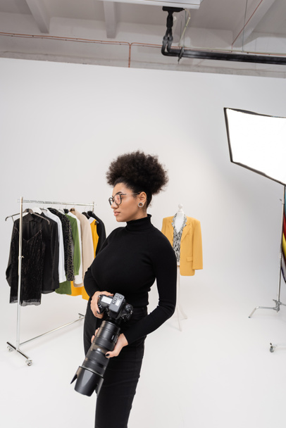 Afrikai amerikai fotós szemüvegben és fekete garbó áll digitális fényképezőgéppel közel gyűjteménye trendi ruhák fotó stúdió - Fotó, kép