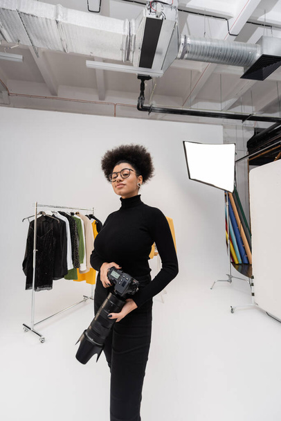 positive african american photographer in eyeglasses standing with digital camera near rail rack with trendy clothes in photo studio - Photo, Image
