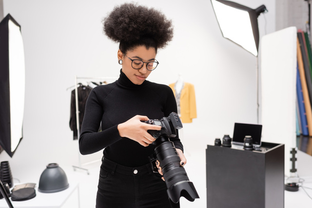 Afrikai amerikai fotós szemüvegben és fekete garbóban nézi a professzionális digitális fényképezőgépet a fotóstúdióban - Fotó, kép