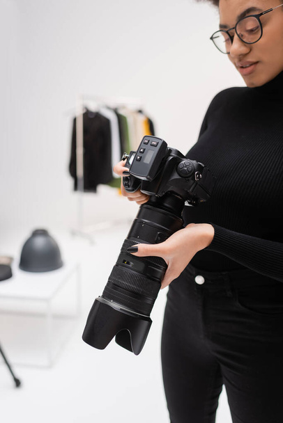 african american content maker in black turtleneck and eyeglasses holding professional digital camera in photo studio - Photo, Image