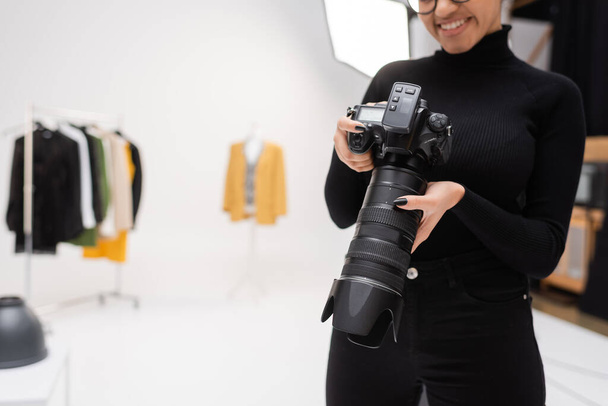 partial view of cheerful african american photographer in black turtleneck holding professional digital camera in blurred photo studio - Photo, image