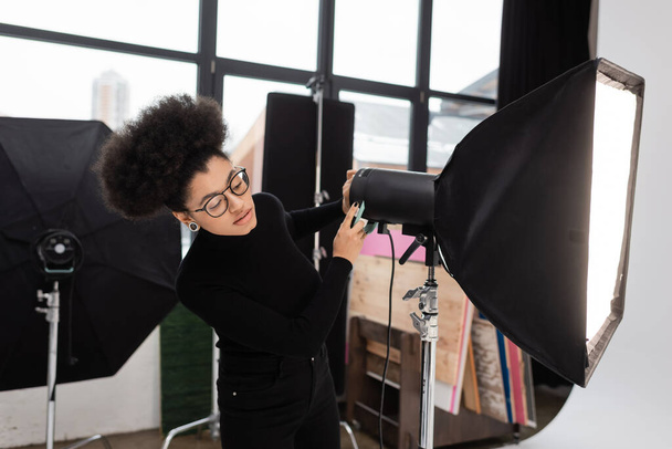 afro-amerikai tartalom producer szemüvegben dolgozik softbox reflektor a fotó stúdióban - Fotó, kép