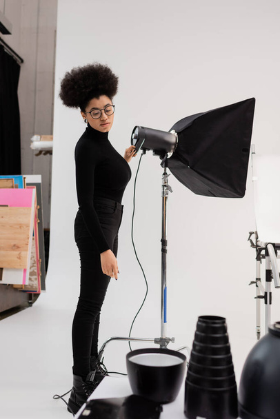 Африканский производитель контента в очках и черной одежде смотрит на световое оборудование в фотостудии - Фото, изображение