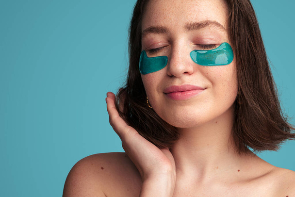 Young sensual female with freckled skin and bare shoulders touching dark hair while having skincare procedure with eye patches against turquoise background - Фото, зображення