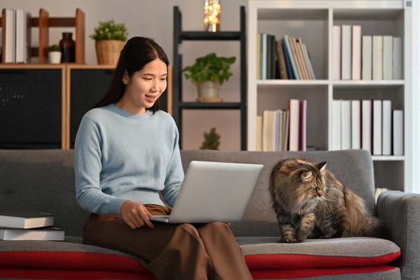 Szczęśliwa młoda kobieta w luźnych ubraniach siedzi na kanapie z szarym puszystym kotem i za pomocą laptopa. - Zdjęcie, obraz