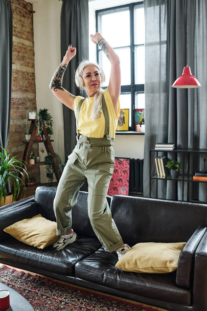 Modern mature Caucasian woman with long gray hair and tattooes on arms listening to music in headphones and dancing on sofa in living room - Фото, зображення