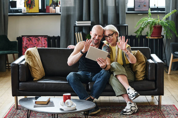 Стильные зрелые мужчина и женщина, сидящие на диване в гостиной приветствуя своих друзей или родственников на видео-звонок на цифровой планшет - Фото, изображение