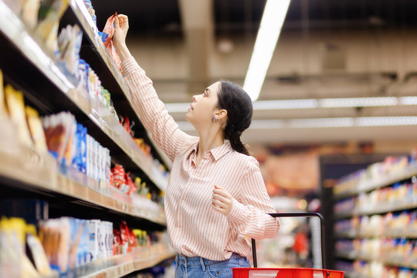Young caucasian housewife takes food from upper shelve. Side view. Showcase at background. Concept of shopping in grocery store and consumerism. - Foto, immagini