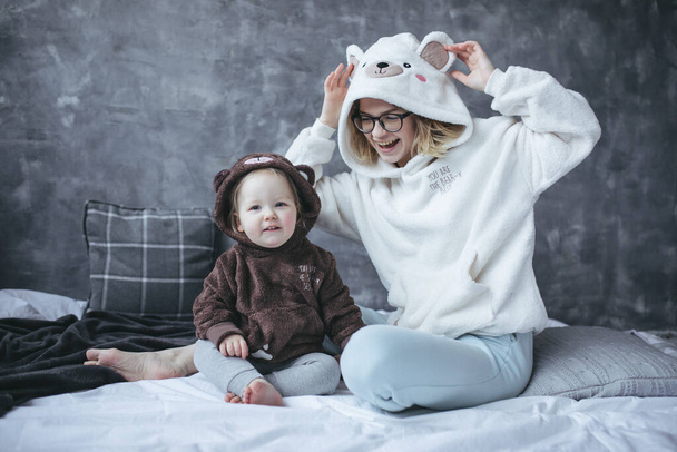 Portrait of family wearing hoodies with bear heads on hoods, sitting on white bed on grey marble background. Young woman mother raising touching ears on hood, playing with daughter. Bedtime, love. - Photo, Image