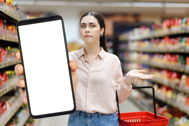 Portrét šokované mladé bělošky drží velký smartphone s bílou obrazovkou a maketou nahoru. Rozumím. Koncept nakupování v obchodě s potravinami. - Fotografie, Obrázek